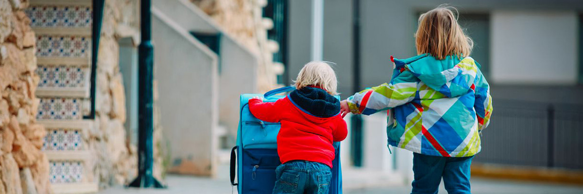 zwei kleine Kinder schieben auf einer Straße einen Koffer, Ansicht von Hinten